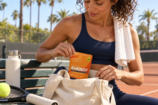 Woman tucking an orange ZYLO Energy packet into a canvas tote beside a tennis court.